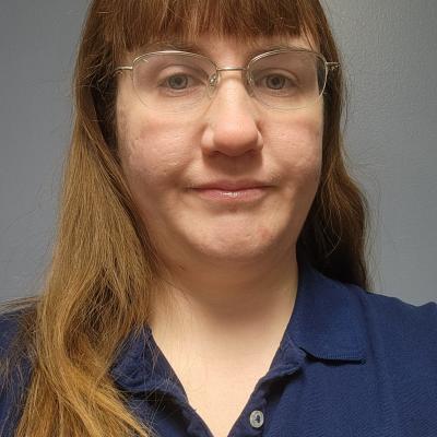 A photo of Christina Kent with long, straight hair and glasses, wearing a navy blue polo shirt, against a simple gray background.