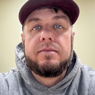 A portrait of William Deming III with a trimmed beard, wearing a maroon baseball cap and a gray hoodie. He gazes directly at the camera against a plain, light background.