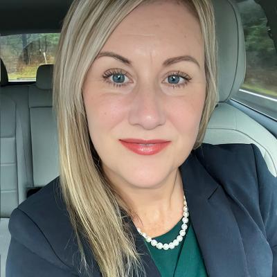 A professional portrait of a woman seated in a car, wearing a dark blazer, a green top, and a pearl necklace. She has light blonde hair and a friendly smile, with a soft outdoor background visible through the car windows.