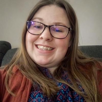 A smiling woman wearing glasses and a patterned top with a rust-colored cardigan takes a photo indoors. The neutral background and her cheerful expression create a warm and approachable vibe.