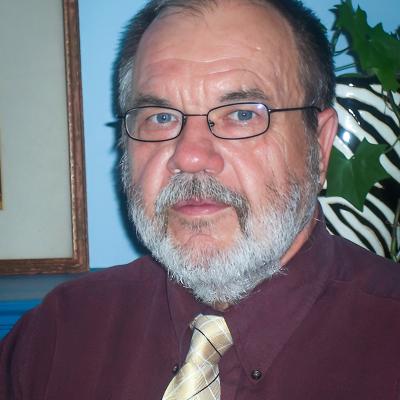 Headshot of Steven Jakobi with glasses and a white beard, wearing a maroon shirt and yellow tie. The background includes a blue wall and greenery.