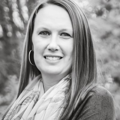 A black-and-white portrait of Jennifer Graves with straight hair, wearing a patterned scarf and hoop earrings. She is smiling warmly, with a natural outdoor backdrop featuring blurred trees.