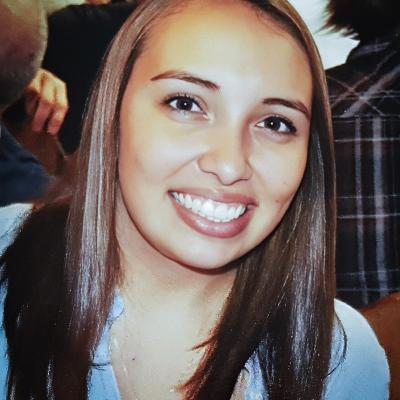 A photo of Cassandra Parker, wearing a light blue shirt and a heart-shaped necklace. The background features a group of people in a casual setting.