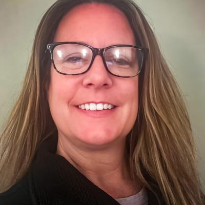 A portrait of Shyla Streb with straight, light brown hair, wearing glasses and a dark zip-up sweater. She smiles brightly against a plain, soft-toned background.