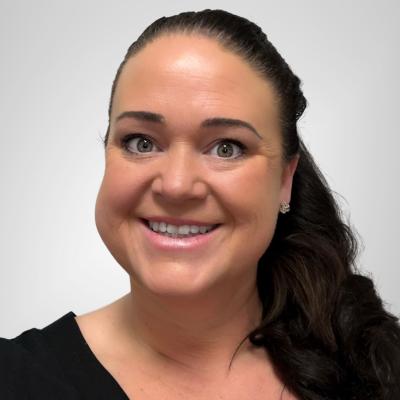 A professional headshot of Lynn Brennan White with long dark hair pulled back, wearing a black top and small stud earrings. She is smiling and set against a plain, light-colored background.