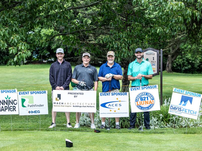 Winning team of the Alfred State golf tournament standing behind tournament sponsor signs