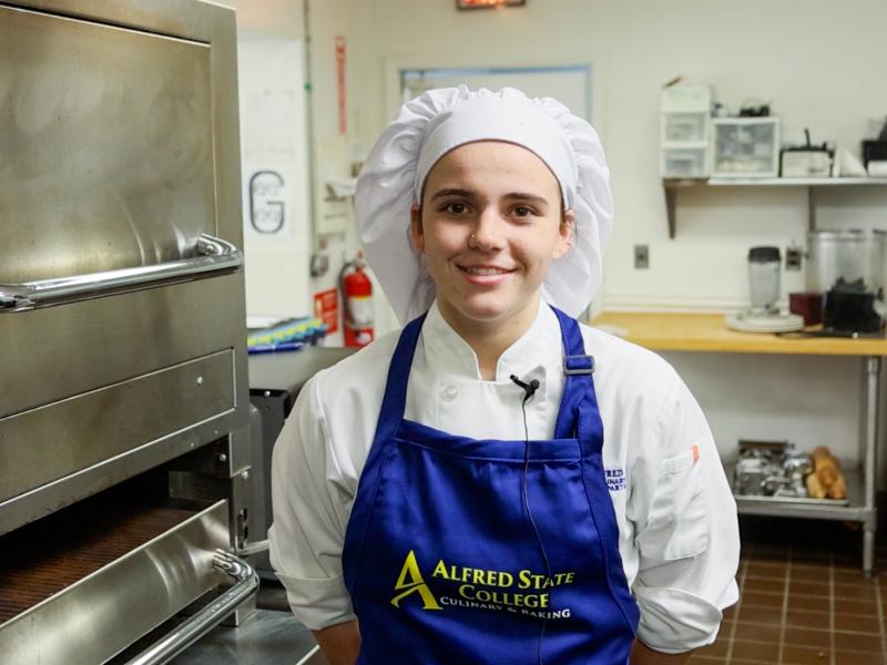 Student poses in a kitchen in front of an oven