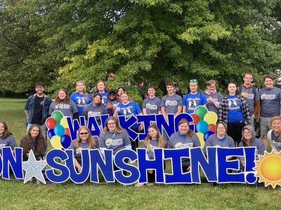 Student stand behind Walking on Sunshine sign