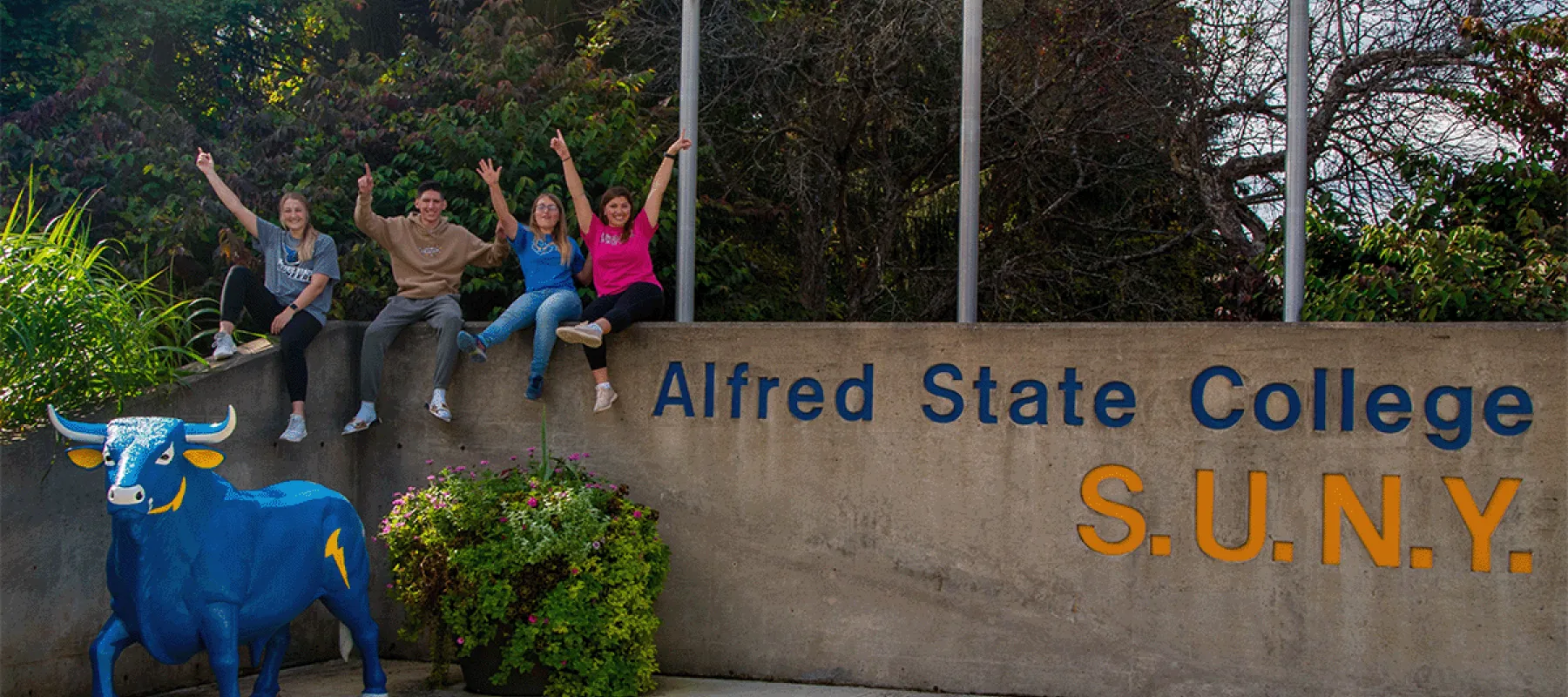 Four students sitting outside at the front entrance to campus with a blue ox and Alfred State College SUNY in view.