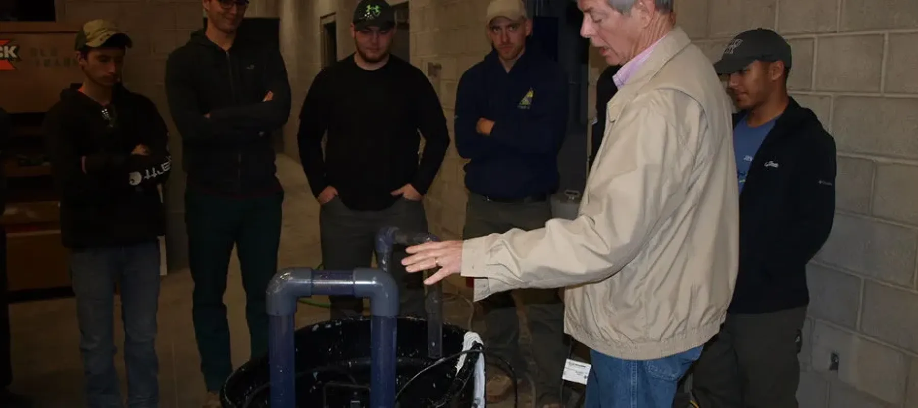 Students gathered around various connected pipes watch demonstration.