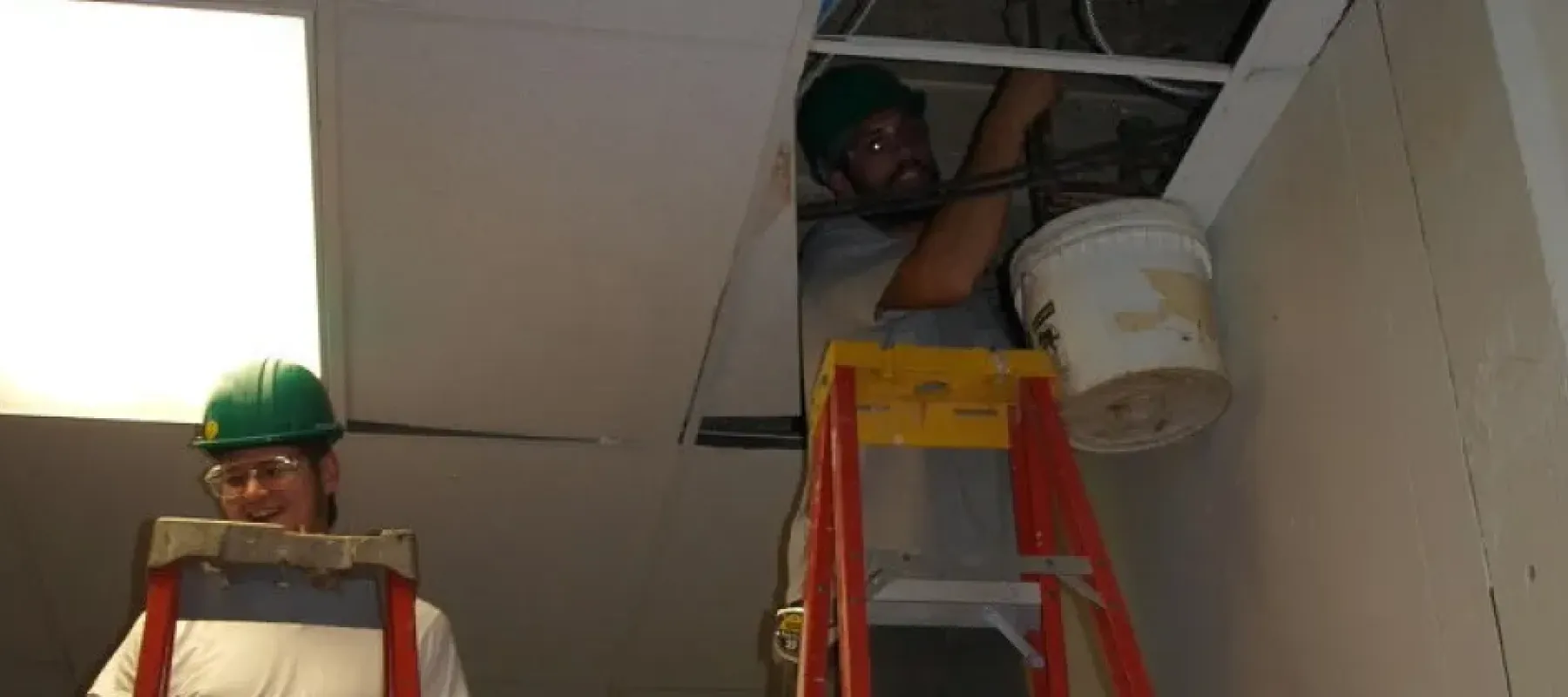 Man on ladder working on pipe inside drop in ceiling.
