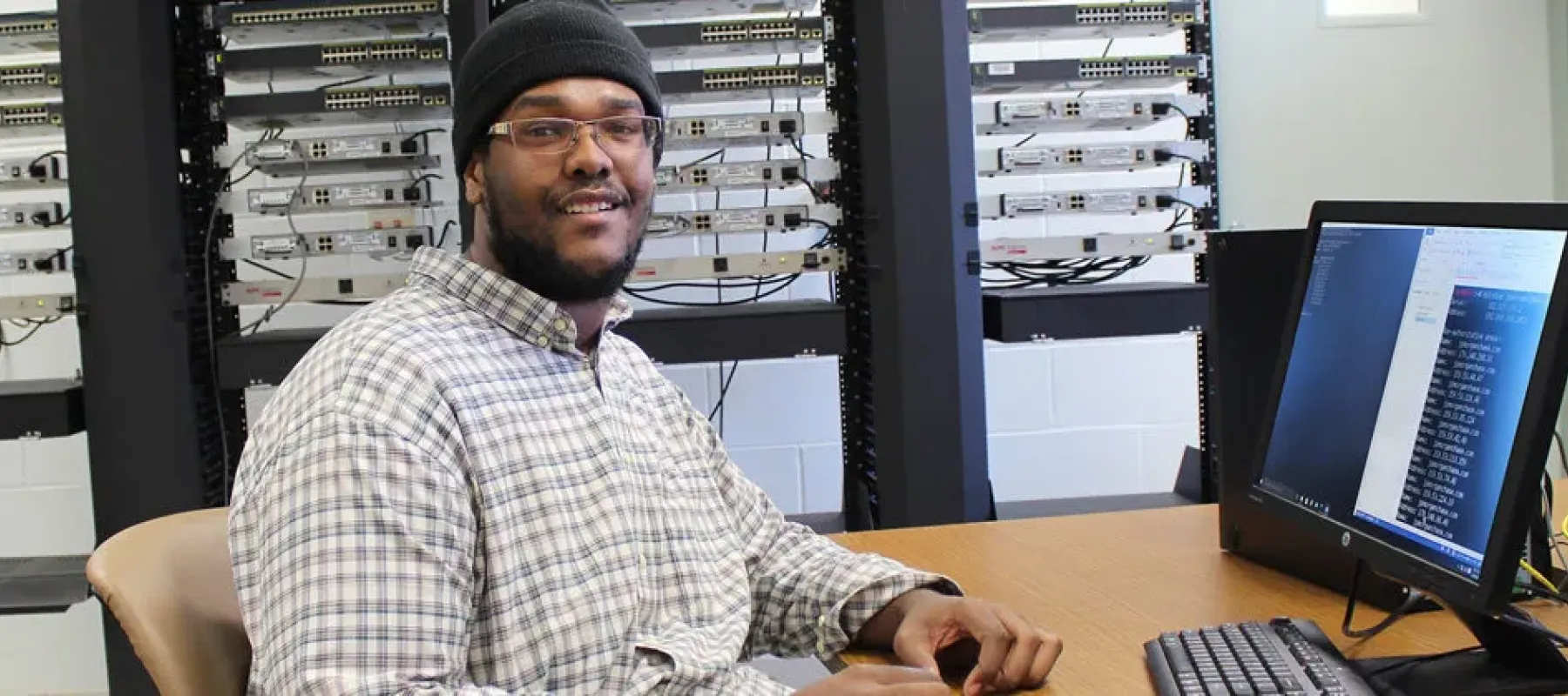 IT student working on computer in classroom with server racks in background.