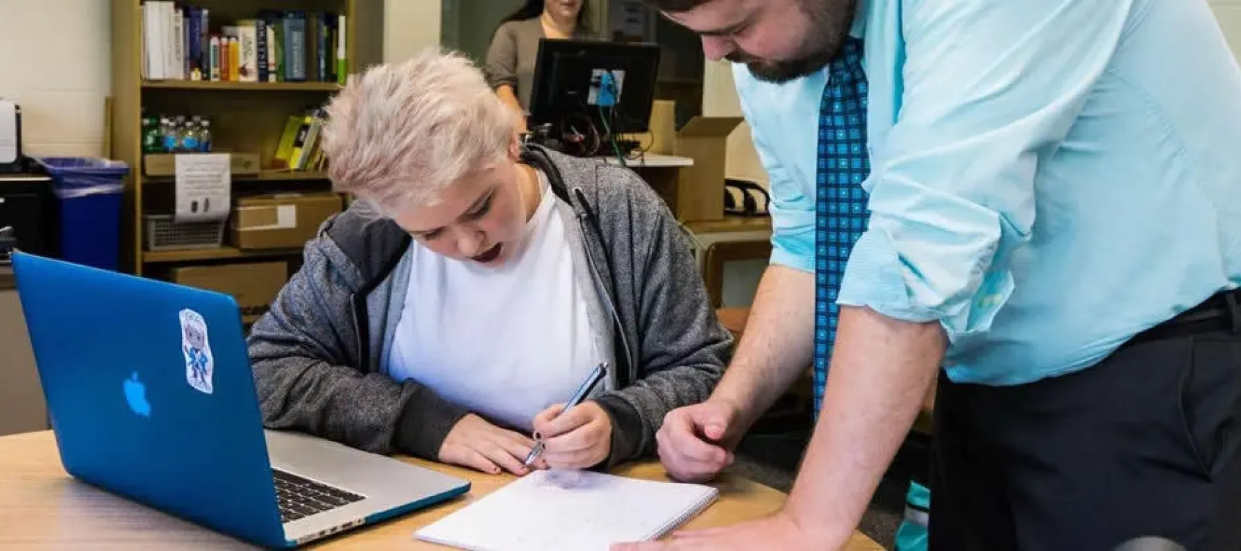 Student sitting at desk receives help with assignment from professor.