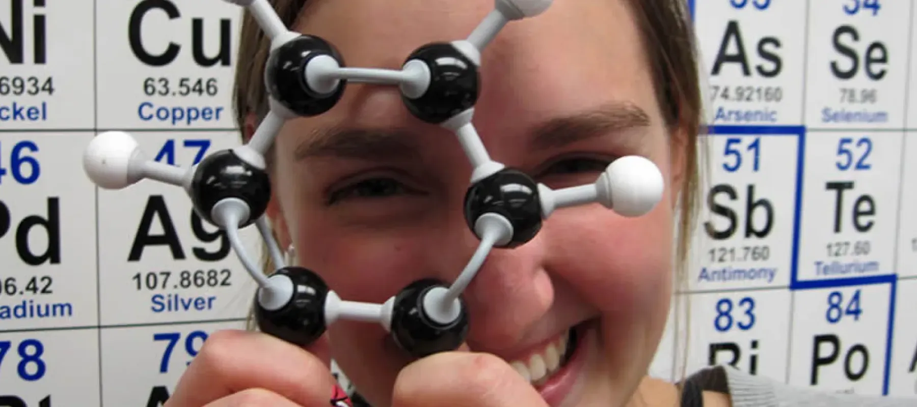 Student holding model of a molecule with periodic table in the background.
