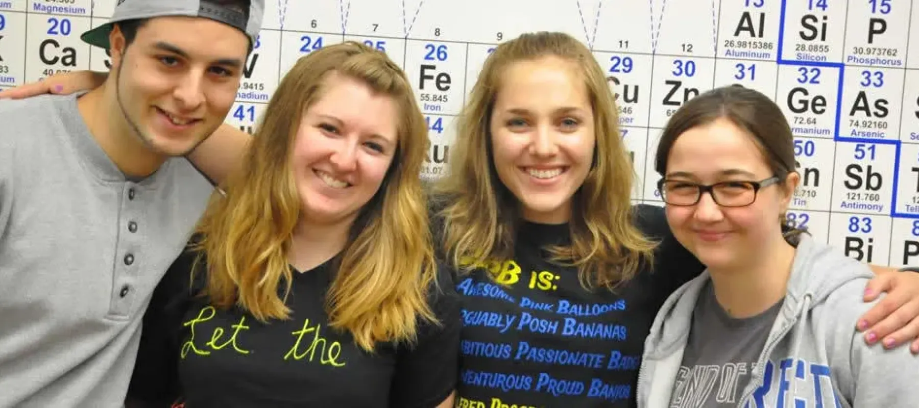 Four students pose in front of periodic table on wall.