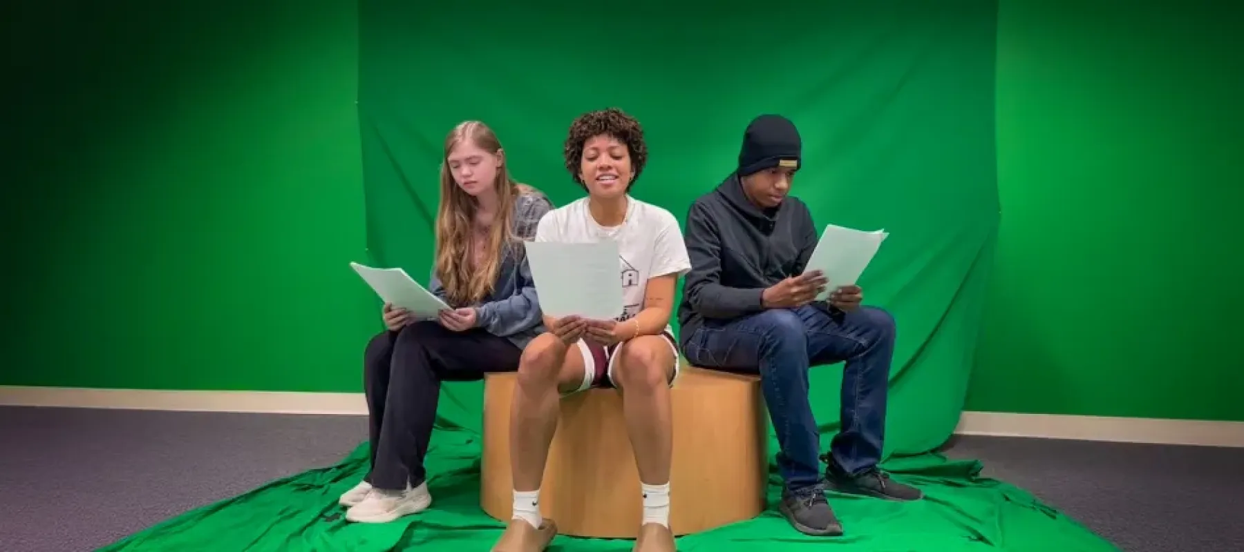Students sitting in front of greenscreen reading from paper.