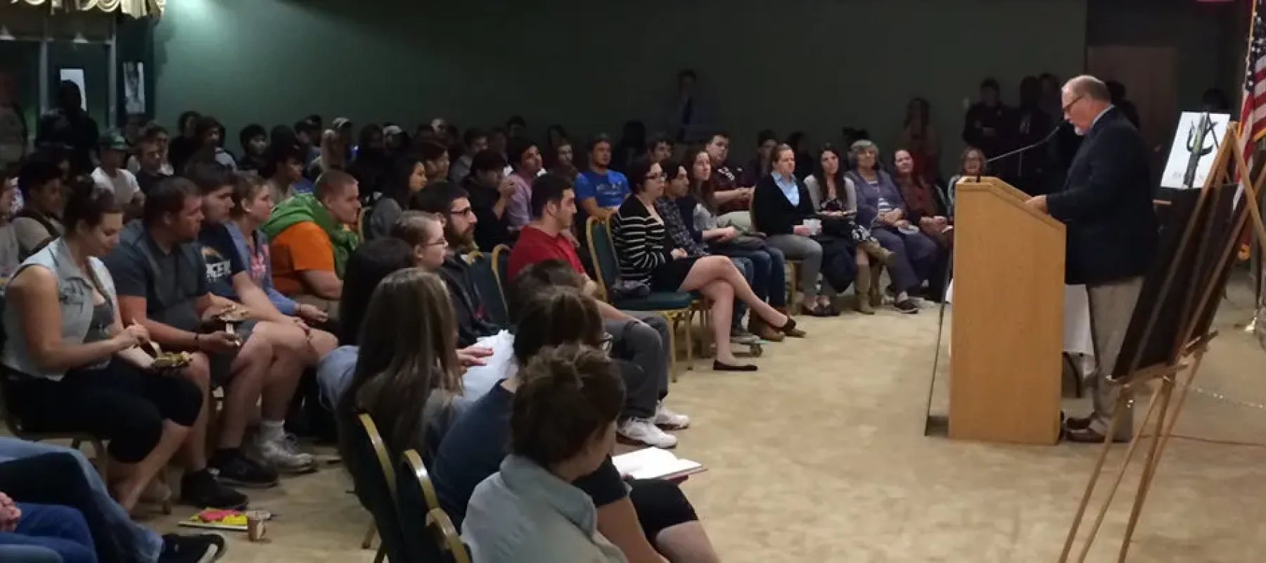 Crowd of people sitting listening to a man standing at a podium speak.