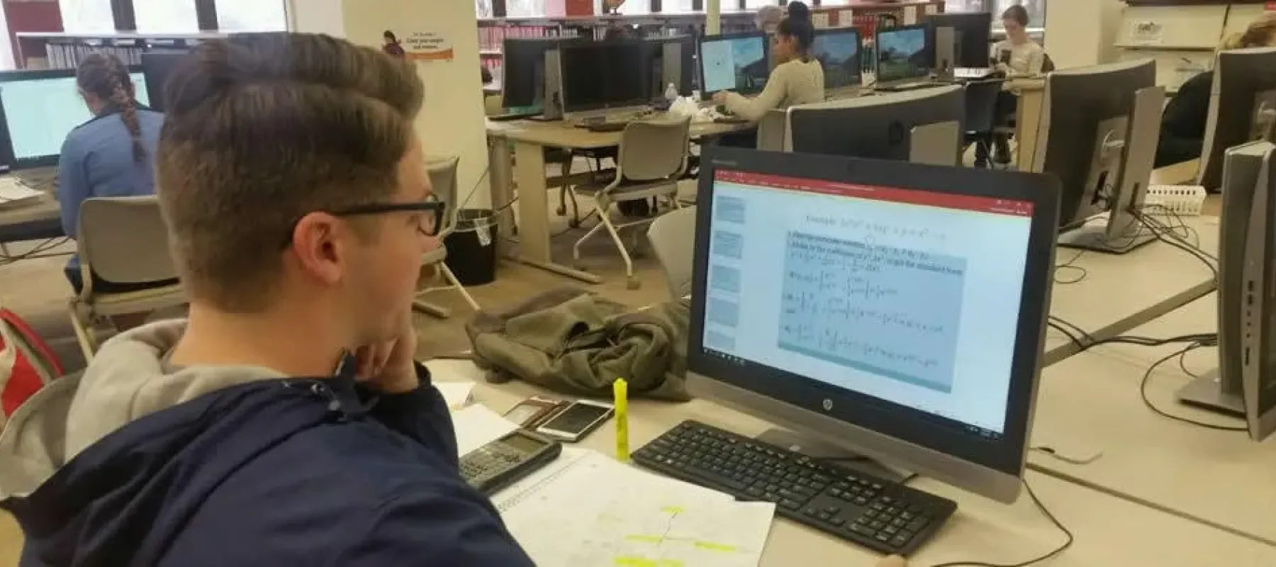 Student sitting at desk with computer showing math equations on a PowerPoint.