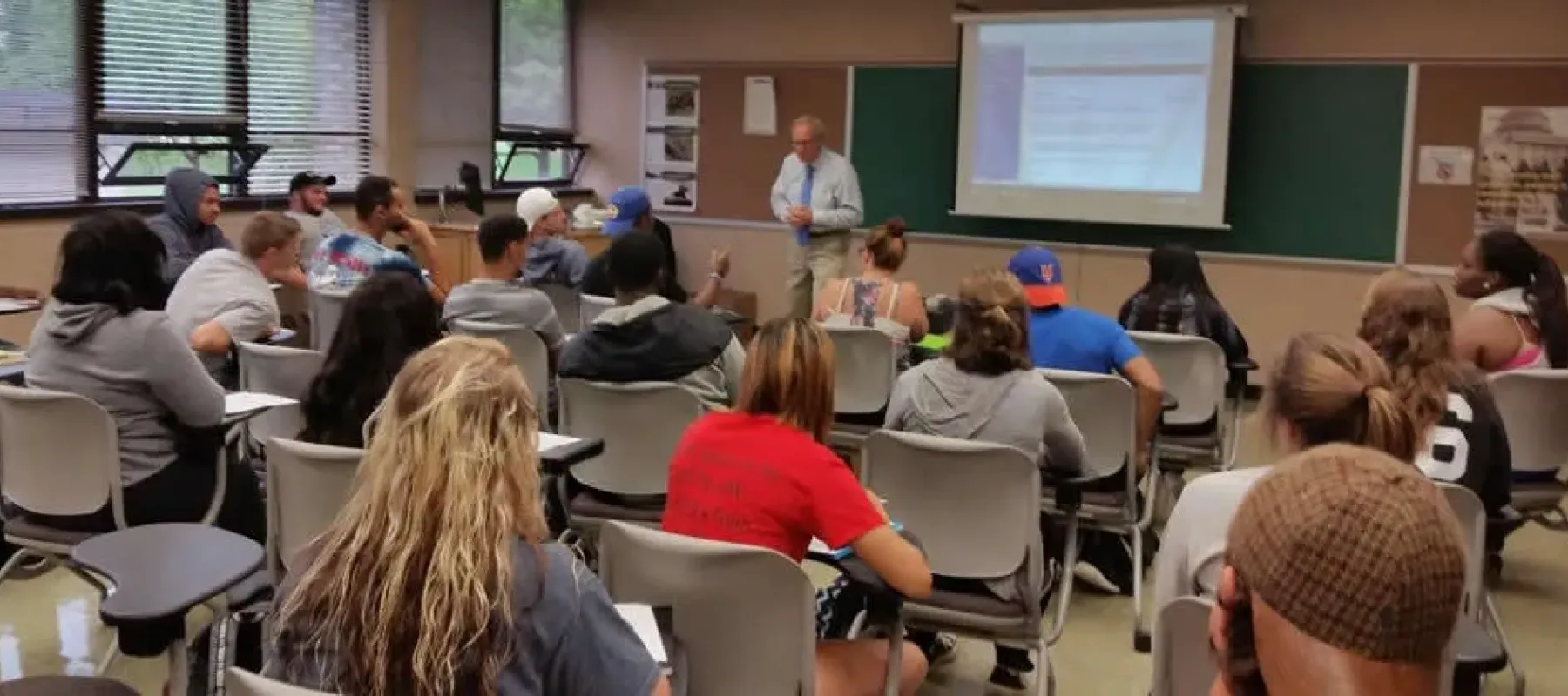 Students sitting as desks in a classroom with the professor in front.