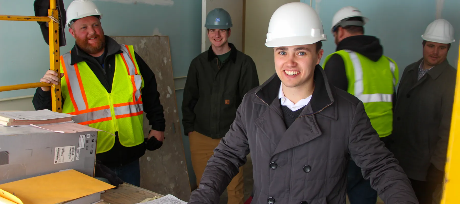 Man with hard hat looking at building plans inside unfished room with other people around.