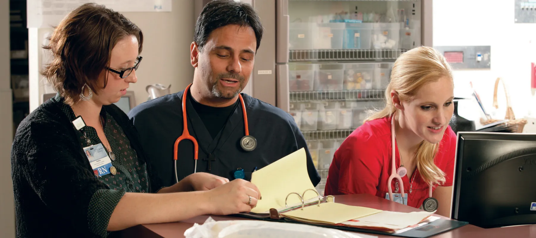 Two medical professionals looking at binder while another is working on a computer in hospital environment.
