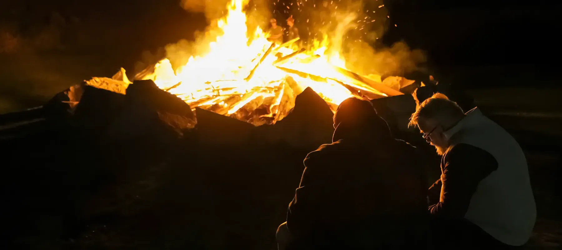 Two people sit near a large bonfire at night, with flames and sparks rising into the sky.
