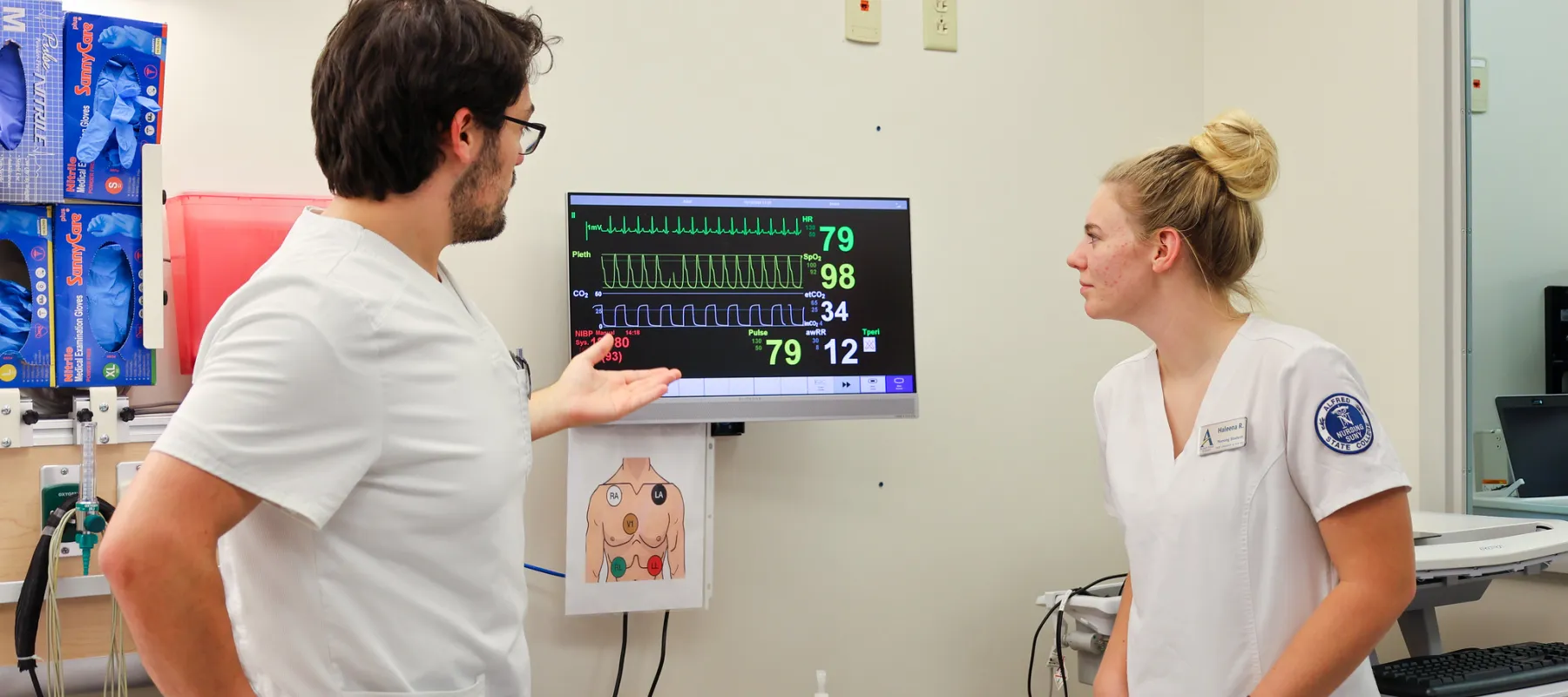 Two nursing students wearing scrubs examining technical equipment in a clinic