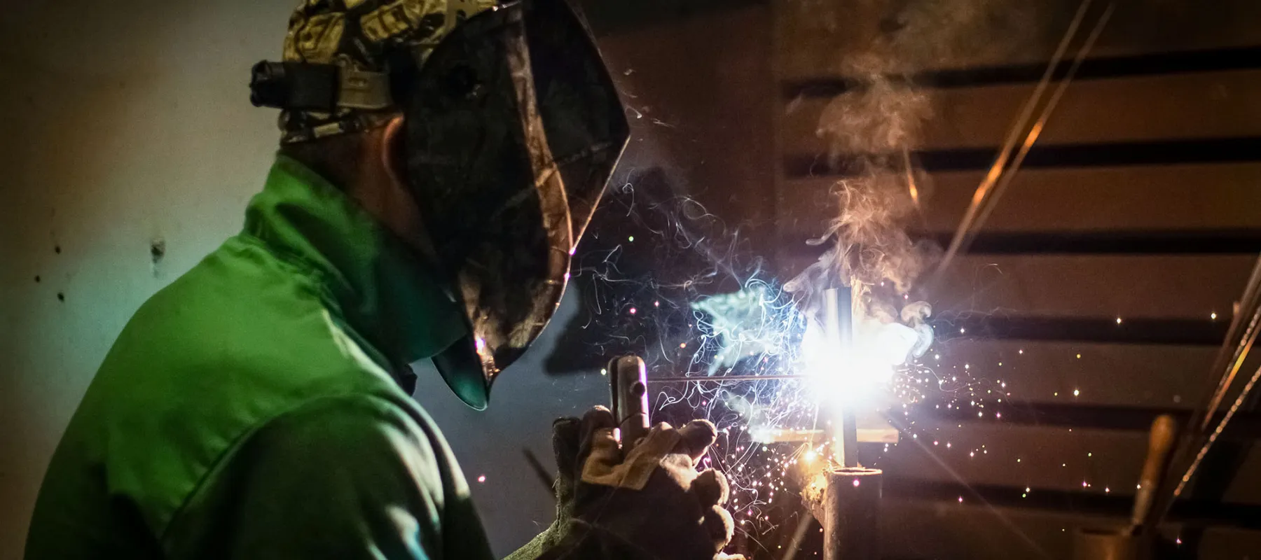 A student wearing safety gear welds, with sparks and smoke rising from the metal.