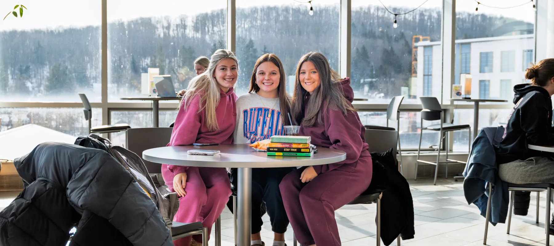 A group of three students sitting around a round table with books and snacks while they smile at the camera.
