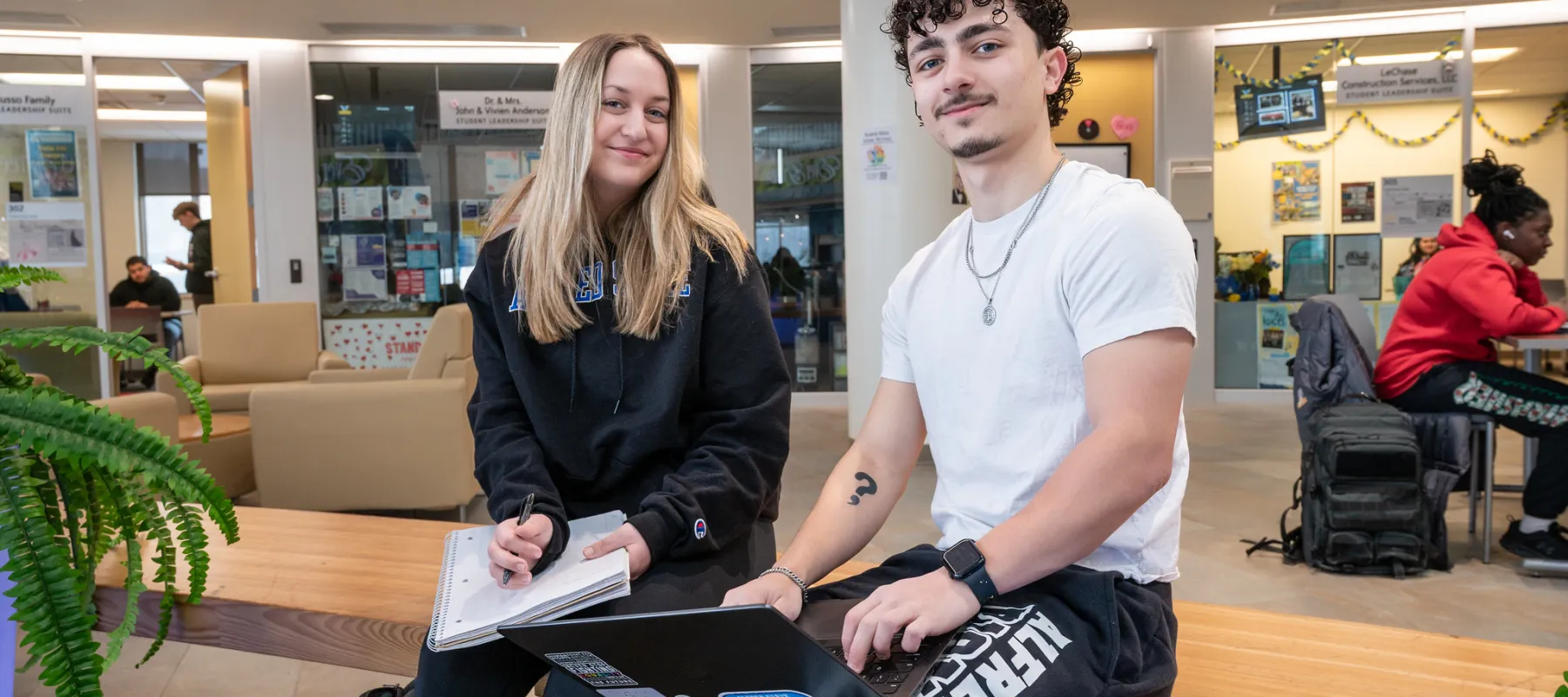Two students sitting on a bench indoors while holding a laptop and notebook.