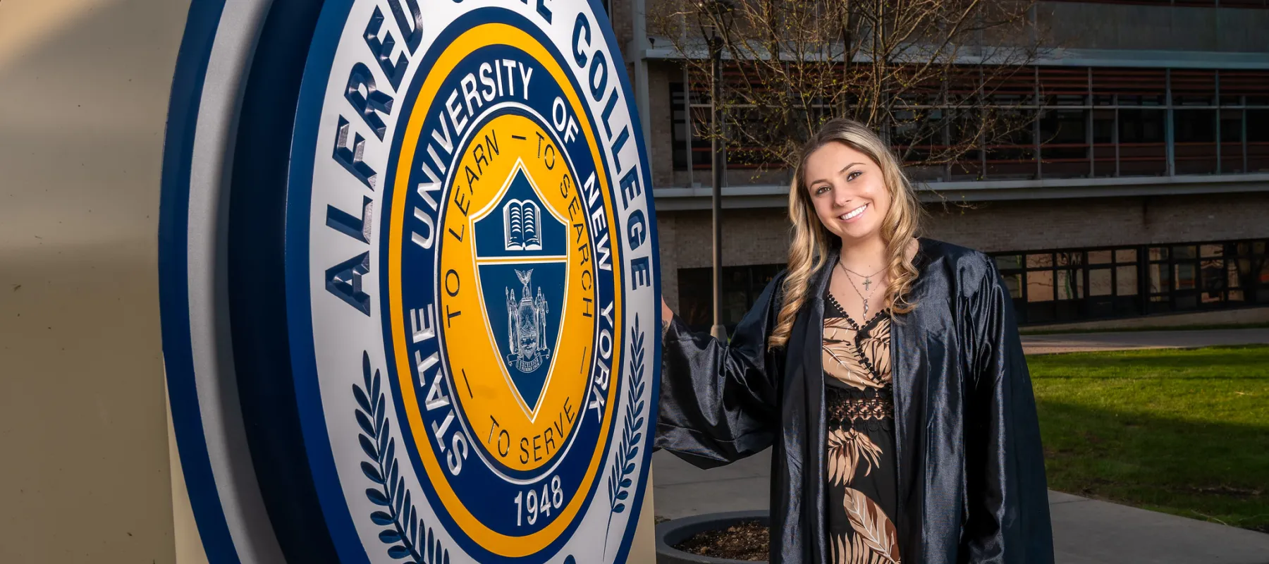 A student wearing a cap and gown standing in front of the Alfred State seal statue.