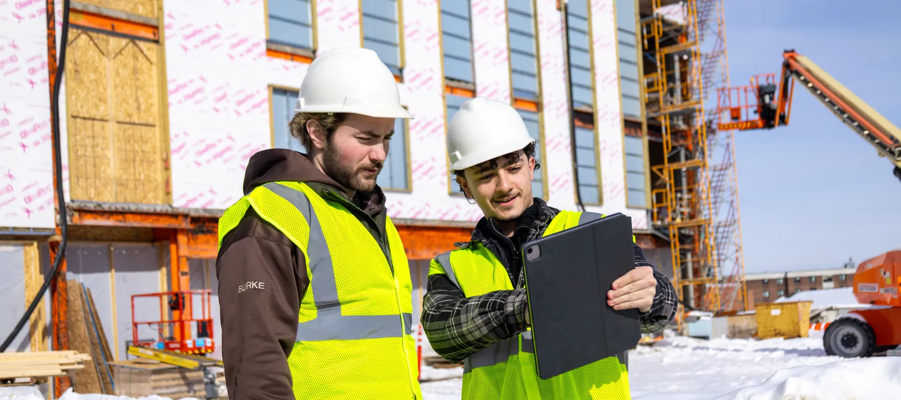 Two students in construction gear reviewing plans on an iPad in a construction site.