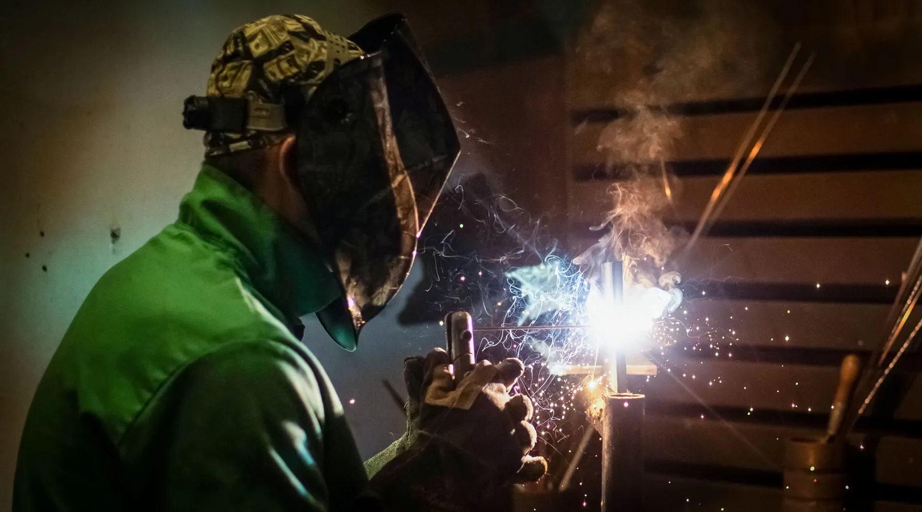 A student wearing safety gear welds, with sparks and smoke rising from the metal.