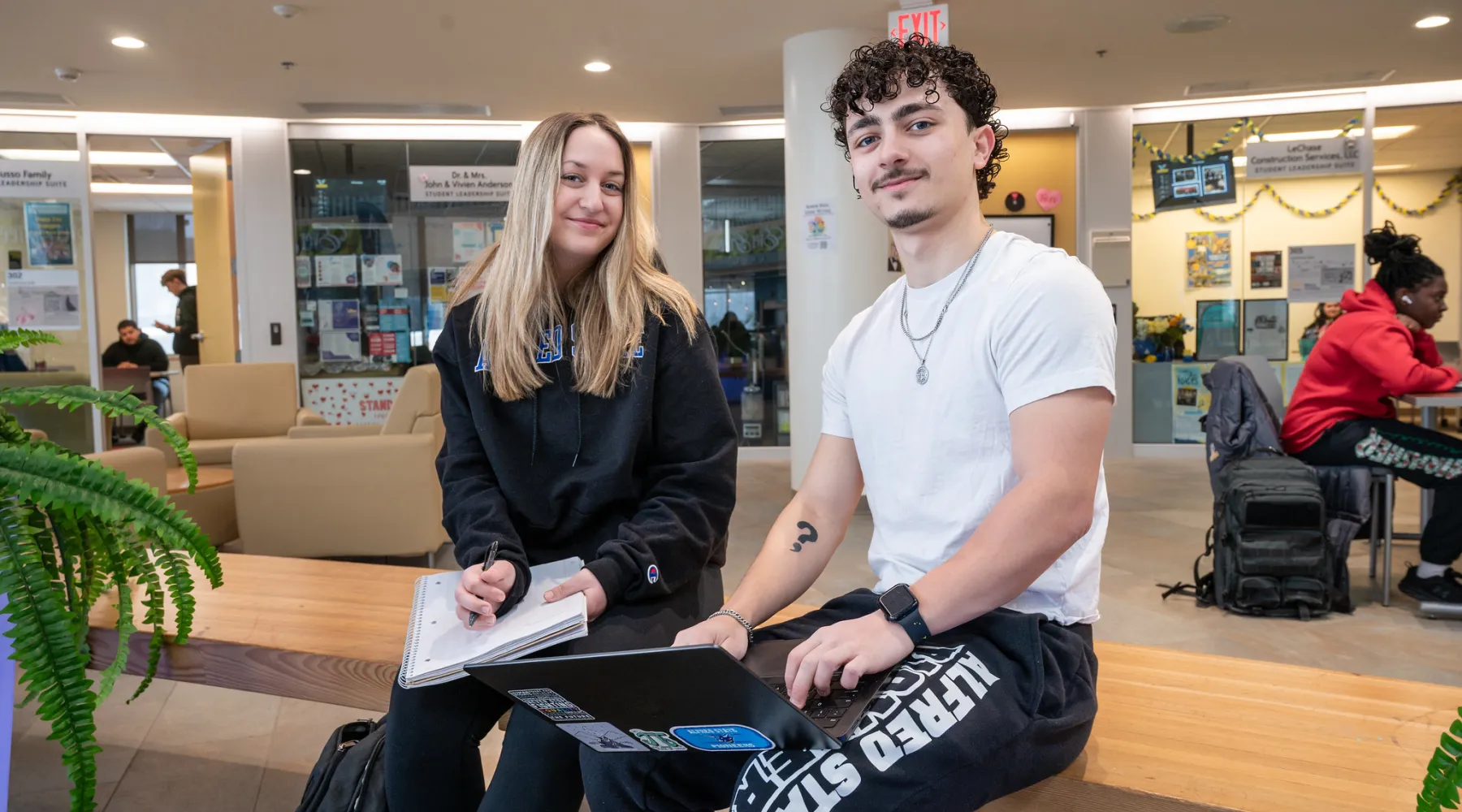 Two students sitting on a bench indoors while holding a laptop and notebook.