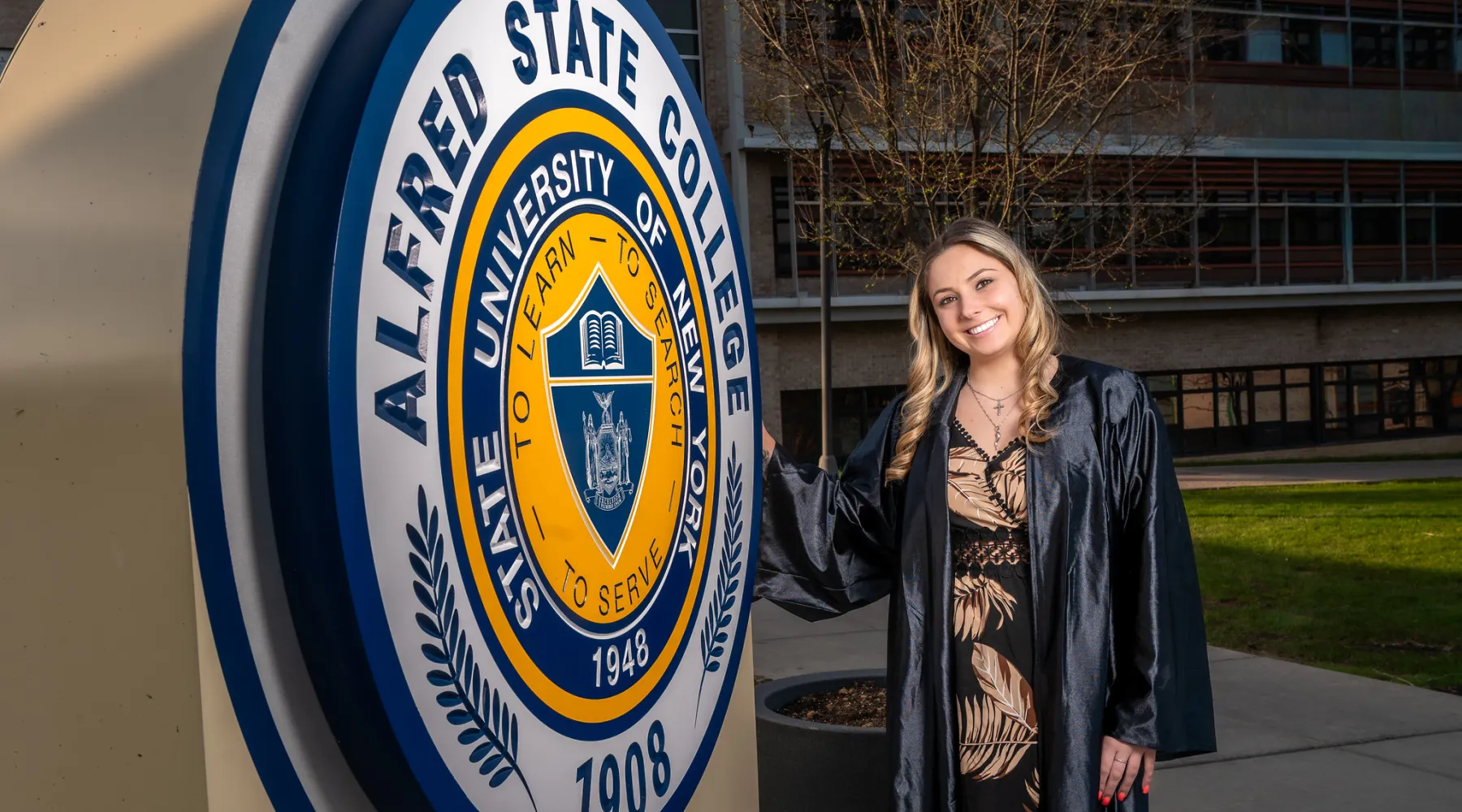 A student wearing a cap and gown standing in front of the Alfred State seal statue.
