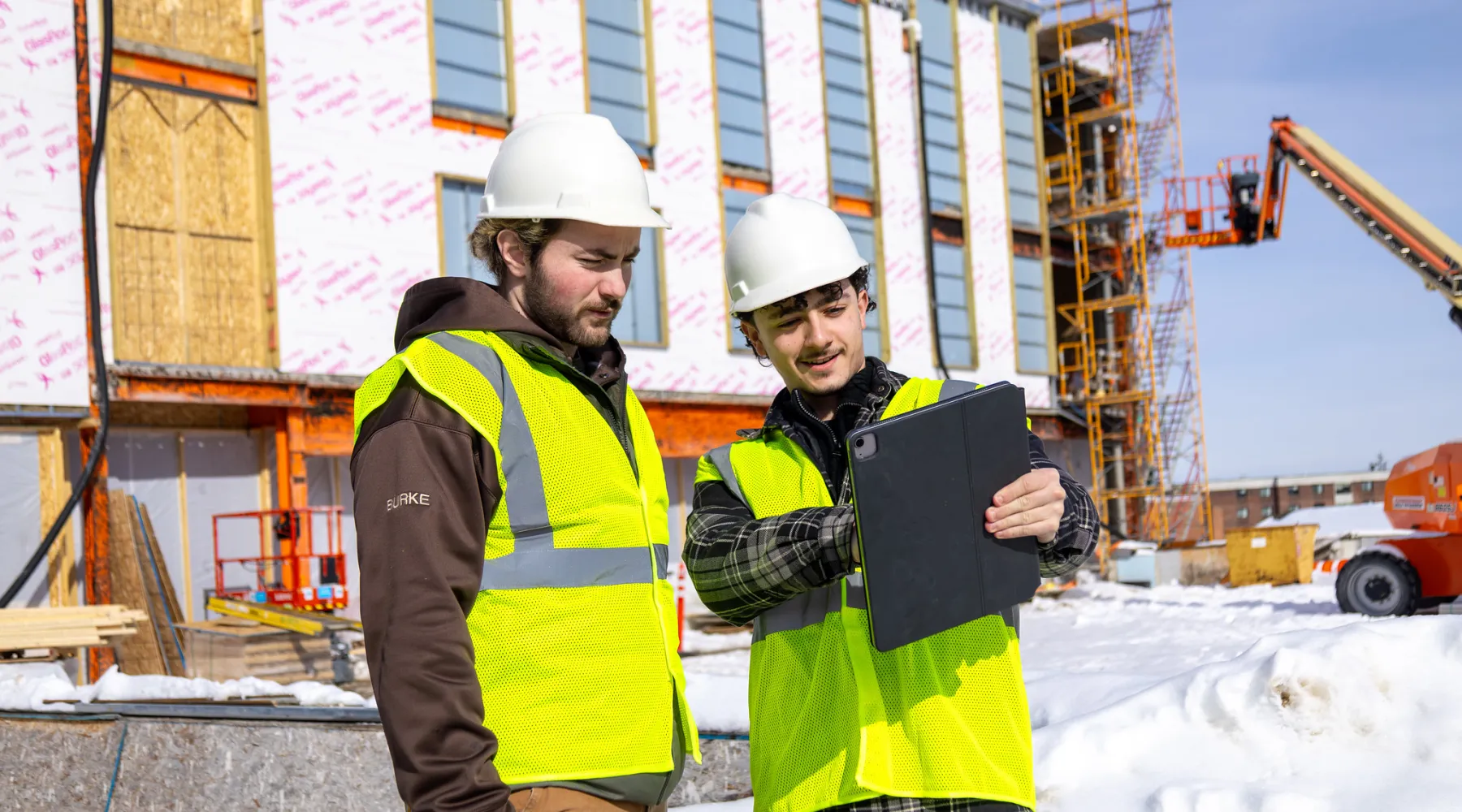Two students in construction gear reviewing plans on an iPad in a construction site.